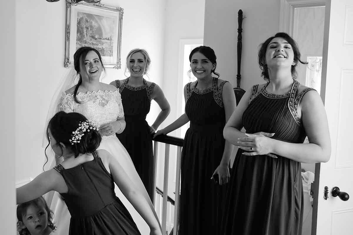 Black and white photo of bride and bridesmaids laughing together on staircase during wedding preparations at Blaithwaite House