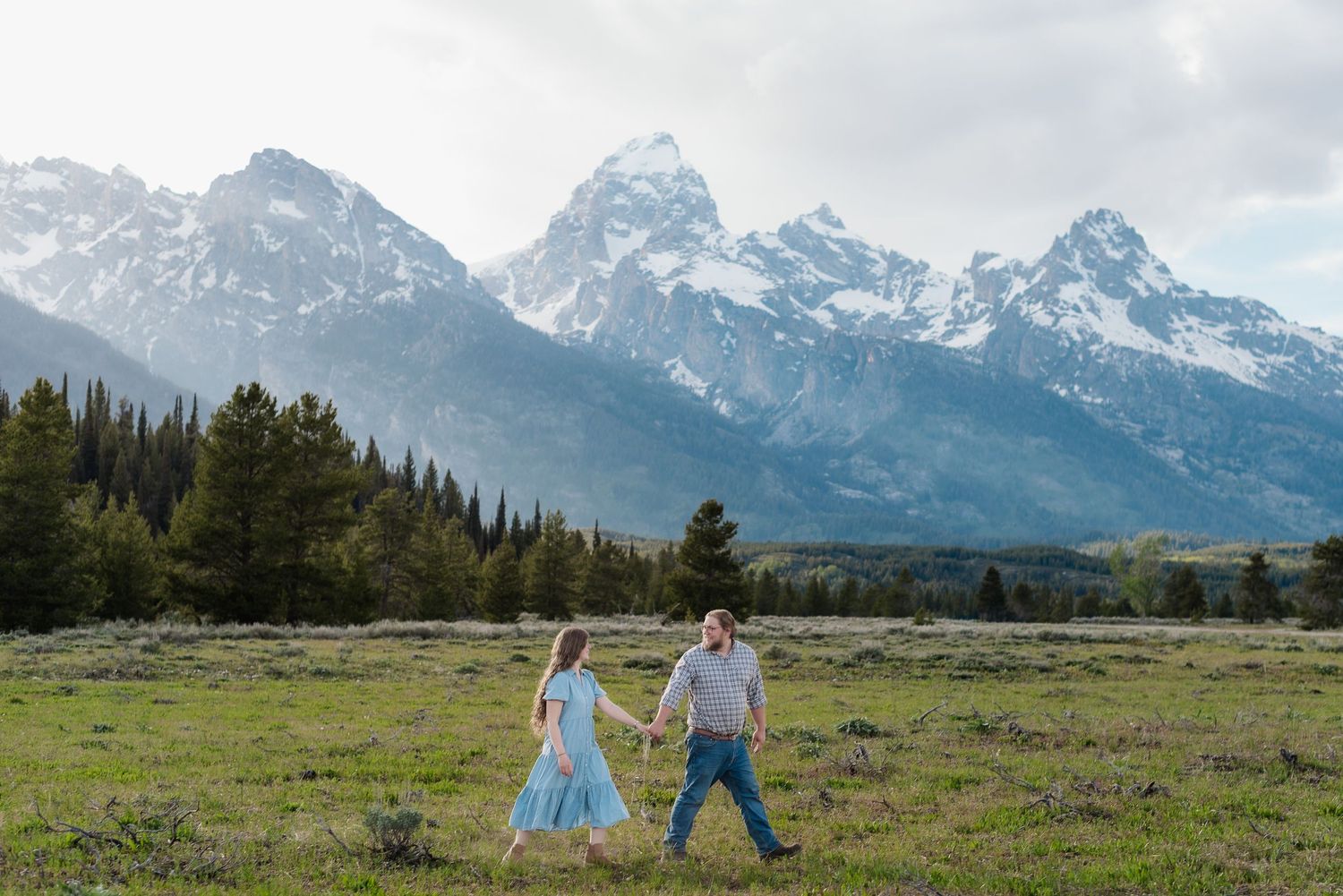 Couple holding hands walking in a green meadow with the snow-capped Grand Teton mountains in the background.