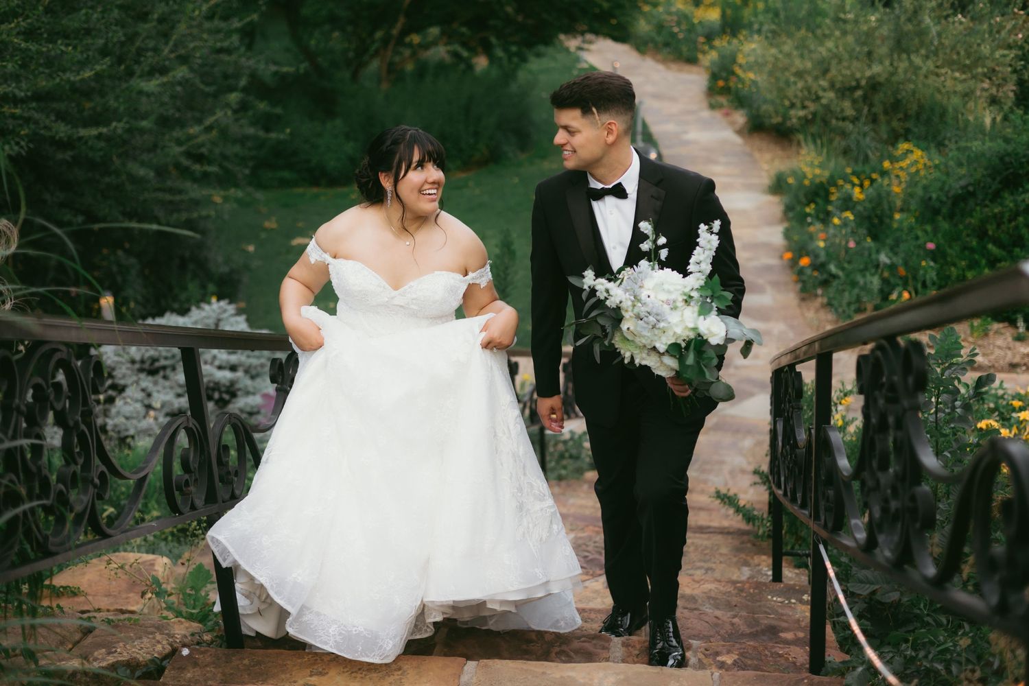 Bride in off-shoulder white gown and groom in black tuxedo walk hand-in-hand down garden stone steps.