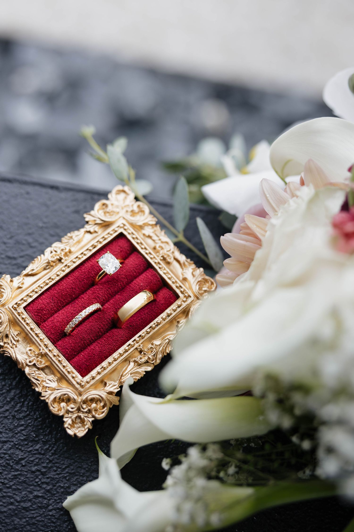 Gold wedding rings with diamond engagement ring displayed in ornate red velvet box beside white floral bouquet.