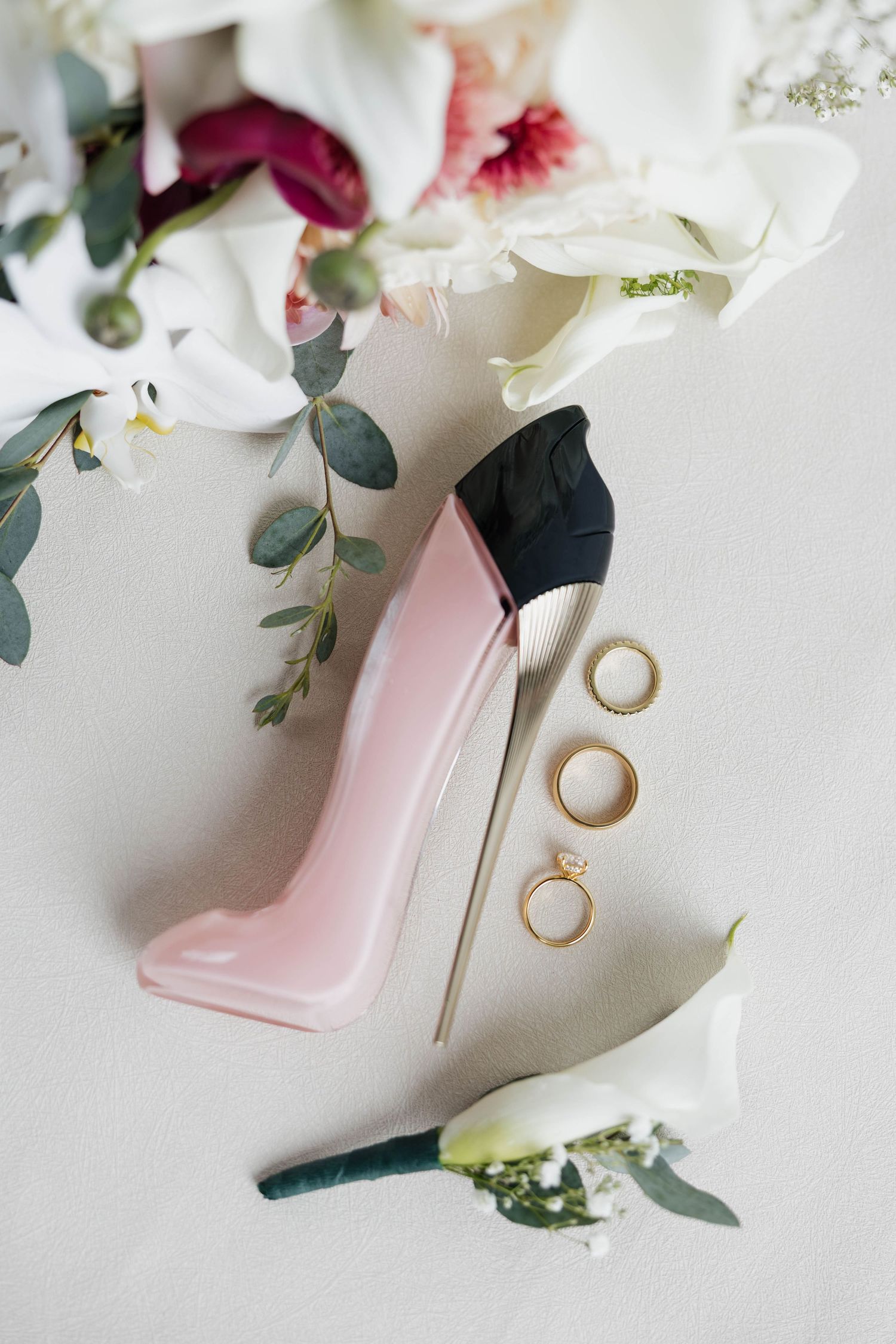 Pink stiletto heel with gold rings and floral bouquet featuring white calla lilies and eucalyptus on a light background.