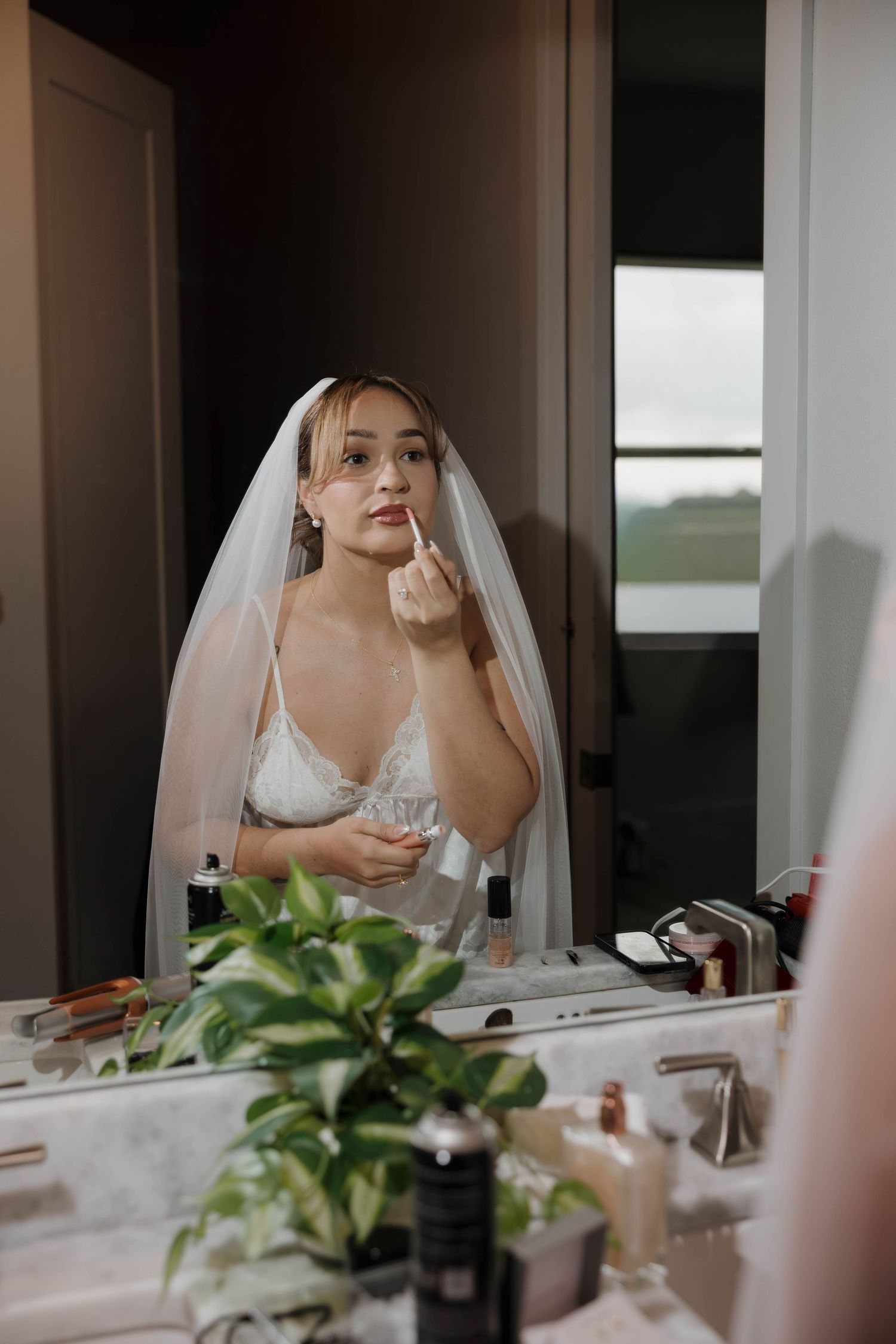 Bride in white lace bra and veil applying makeup at vanity mirror on wedding day.