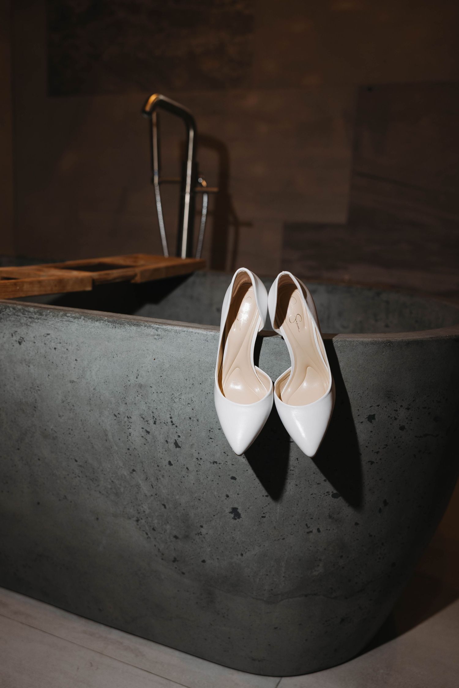 White bridal heels displayed on the edge of a modern concrete bathtub in a dark, moody bathroom setting.