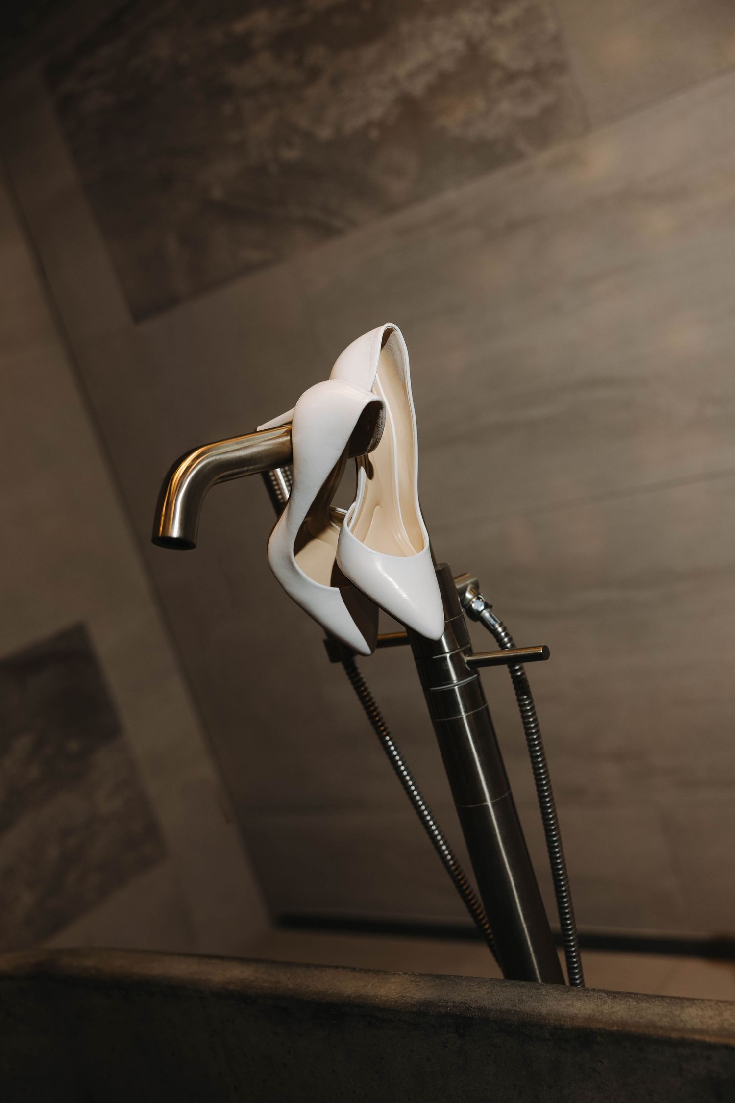 White bridal heels balanced on a gold bathroom faucet and fixtures against a marble wall backdrop.
