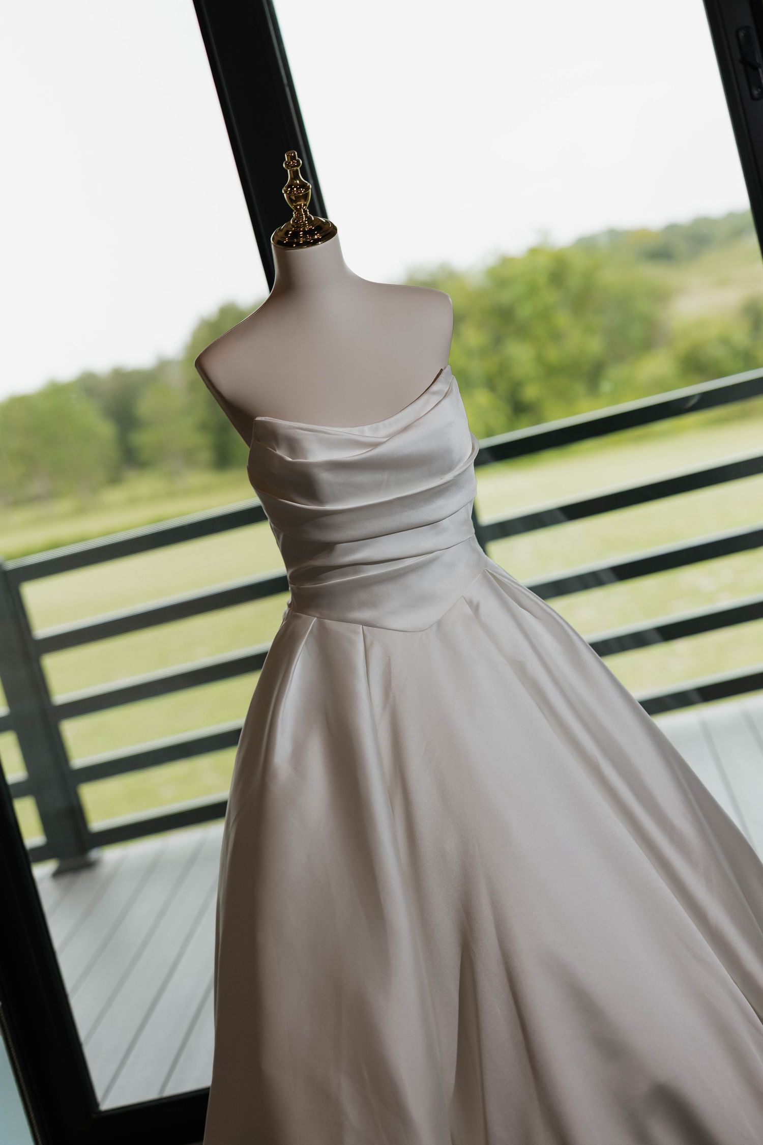 Elegant strapless satin wedding gown displayed on a mannequin near large windows with a lush green landscape backdrop.