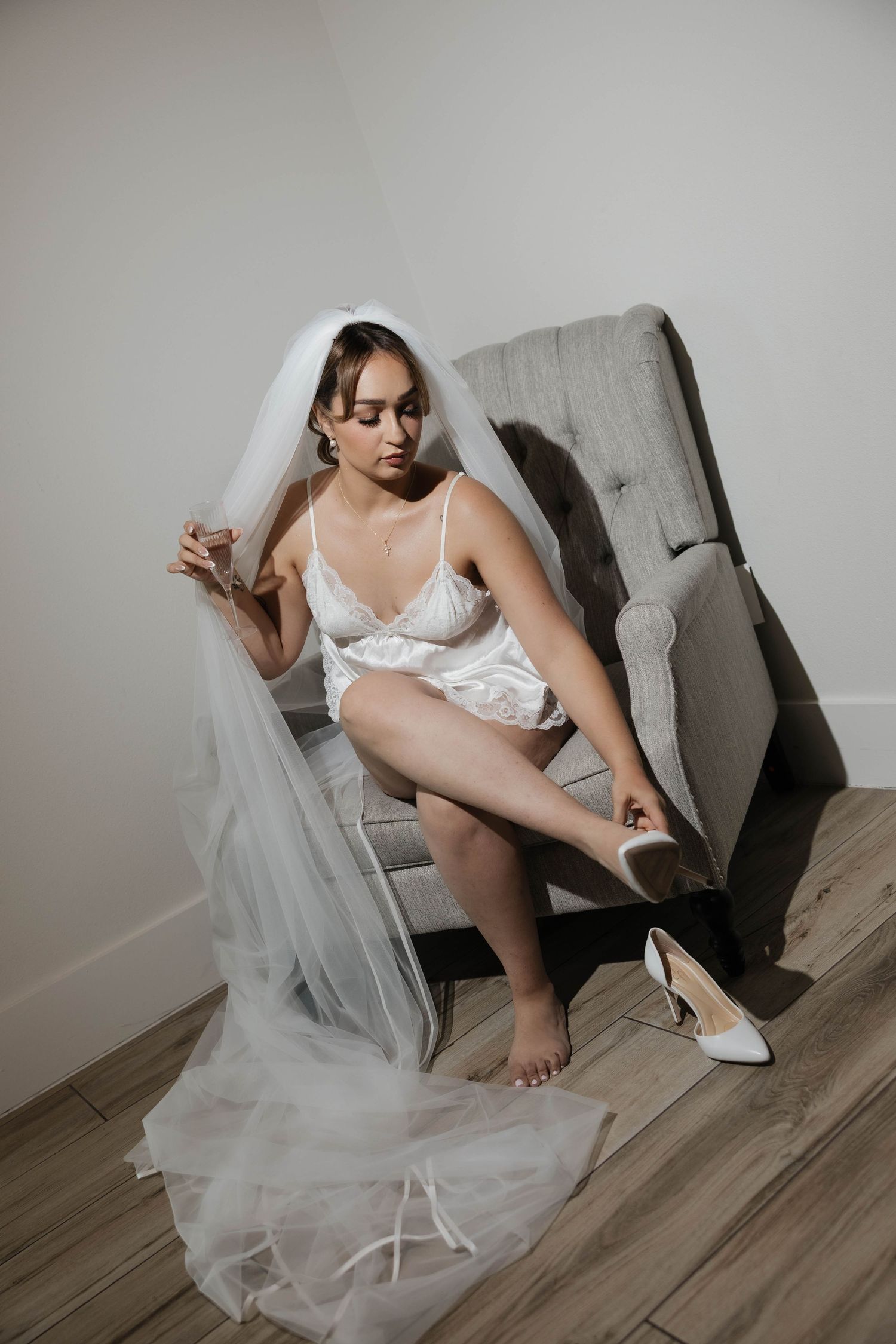 Bride in white lace lingerie and veil sits in gray chair, putting on white heels before her wedding ceremony.