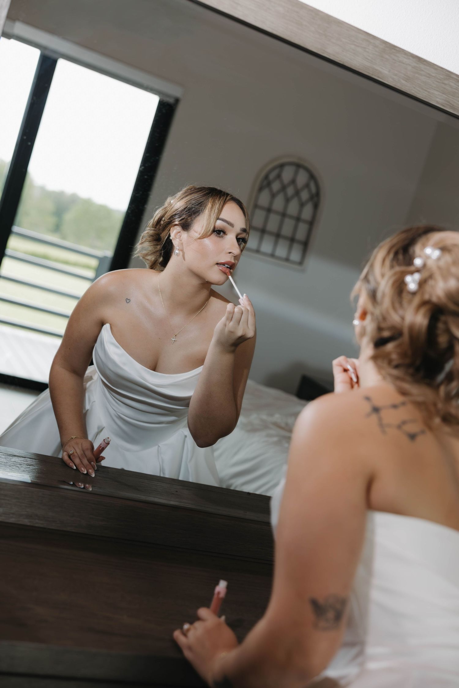 Bride in white strapless gown applying lipstick while gazing into mirror during wedding day preparations.