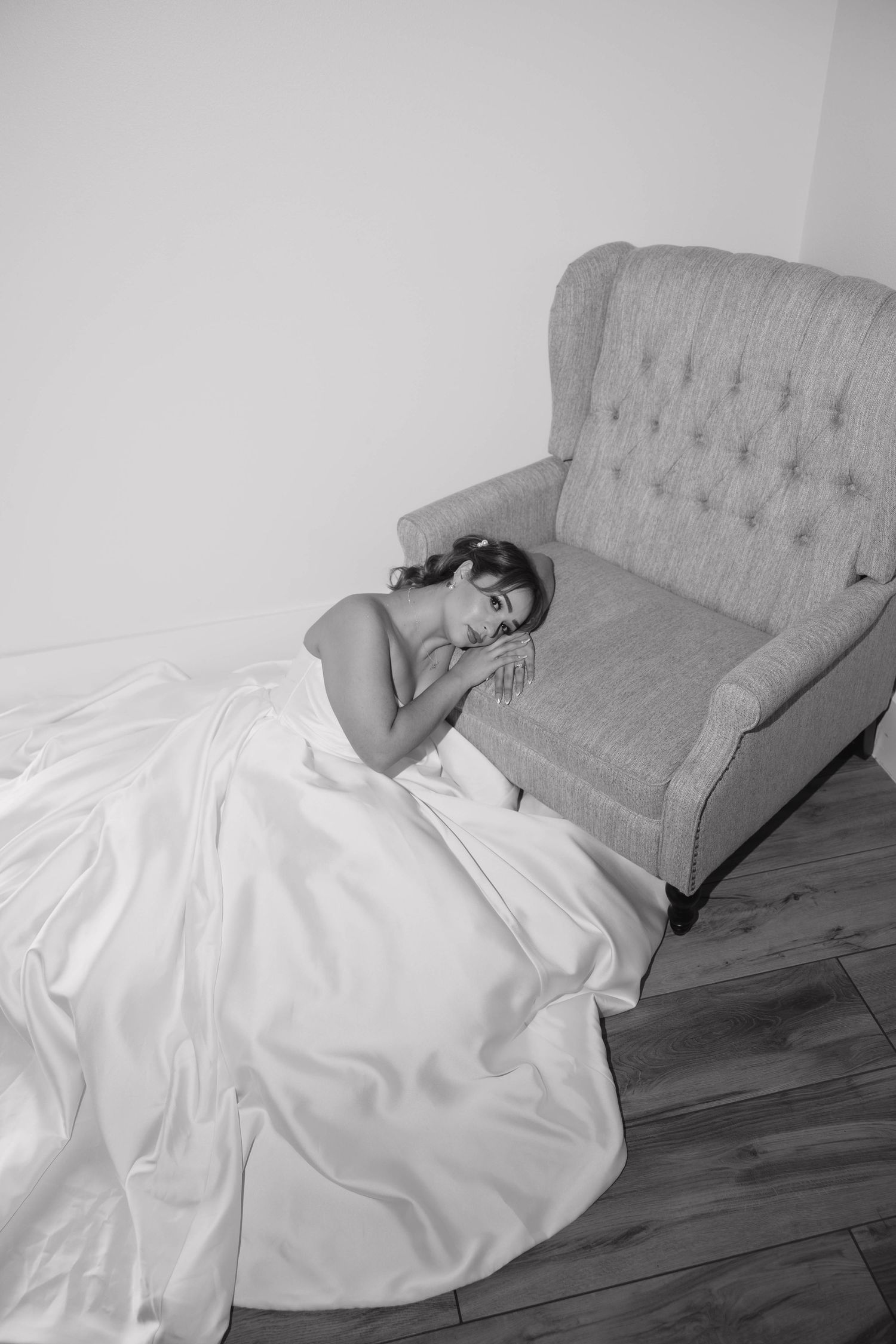 Black and white photo of a bride in a flowing white gown resting on a tufted armchair on a hardwood floor.