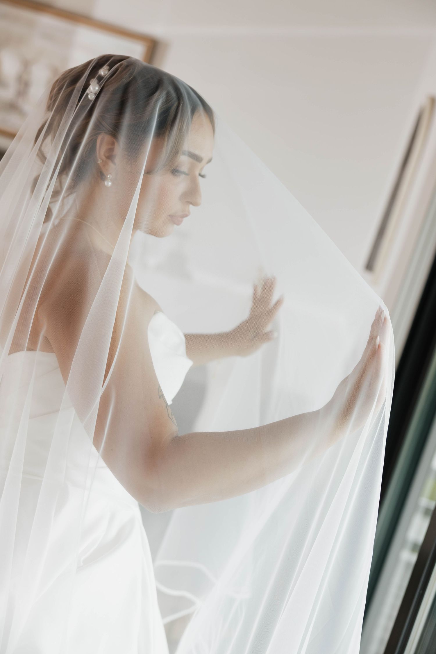 Bride in white gown and long veil looking down gracefully near a window on her wedding day.