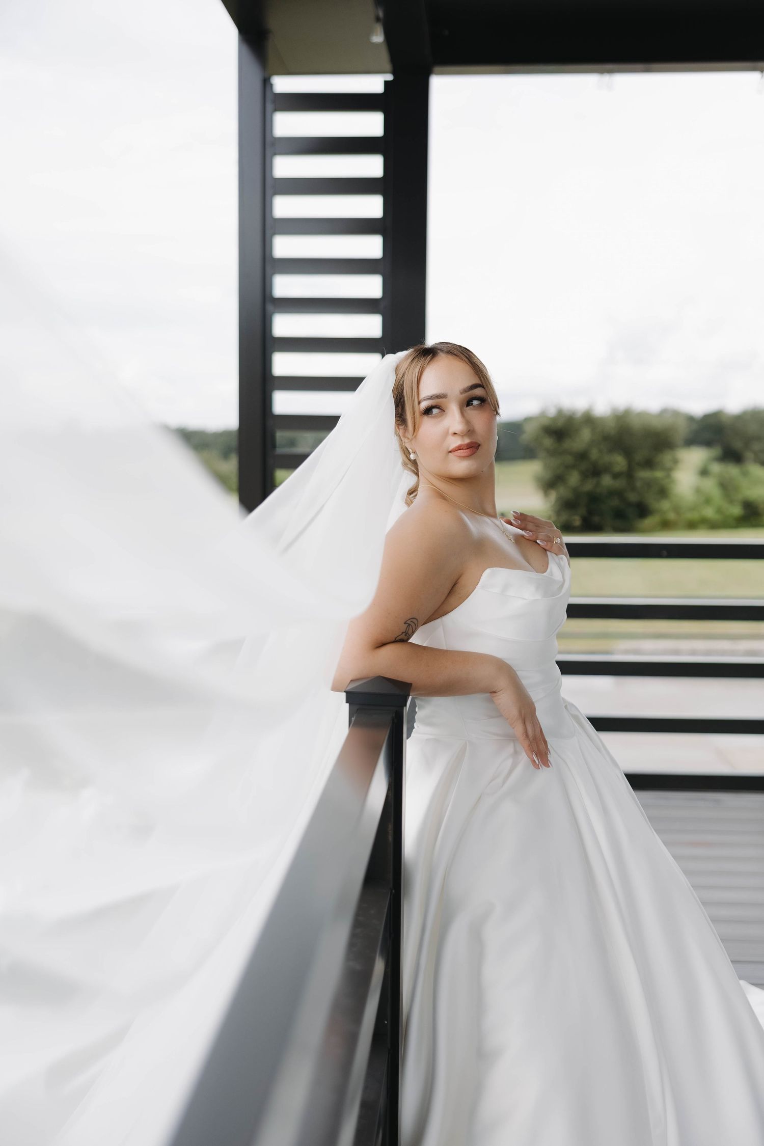 Elegant bride in white ball gown and veil poses on balcony with scenic green landscape backdrop.