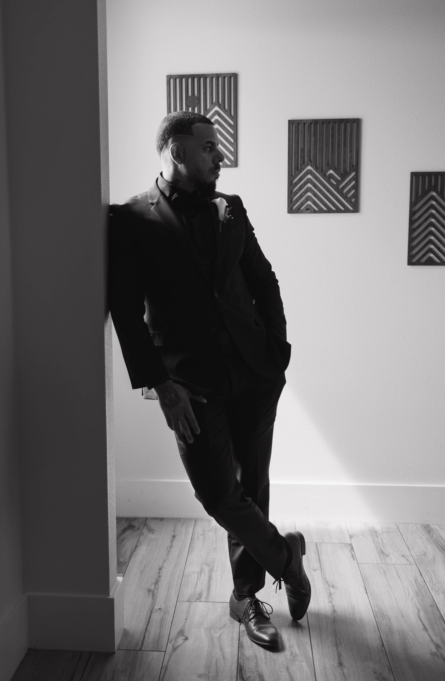 Black and white photo of a stylish man in a dark suit leaning against a white wall with geometric art prints.