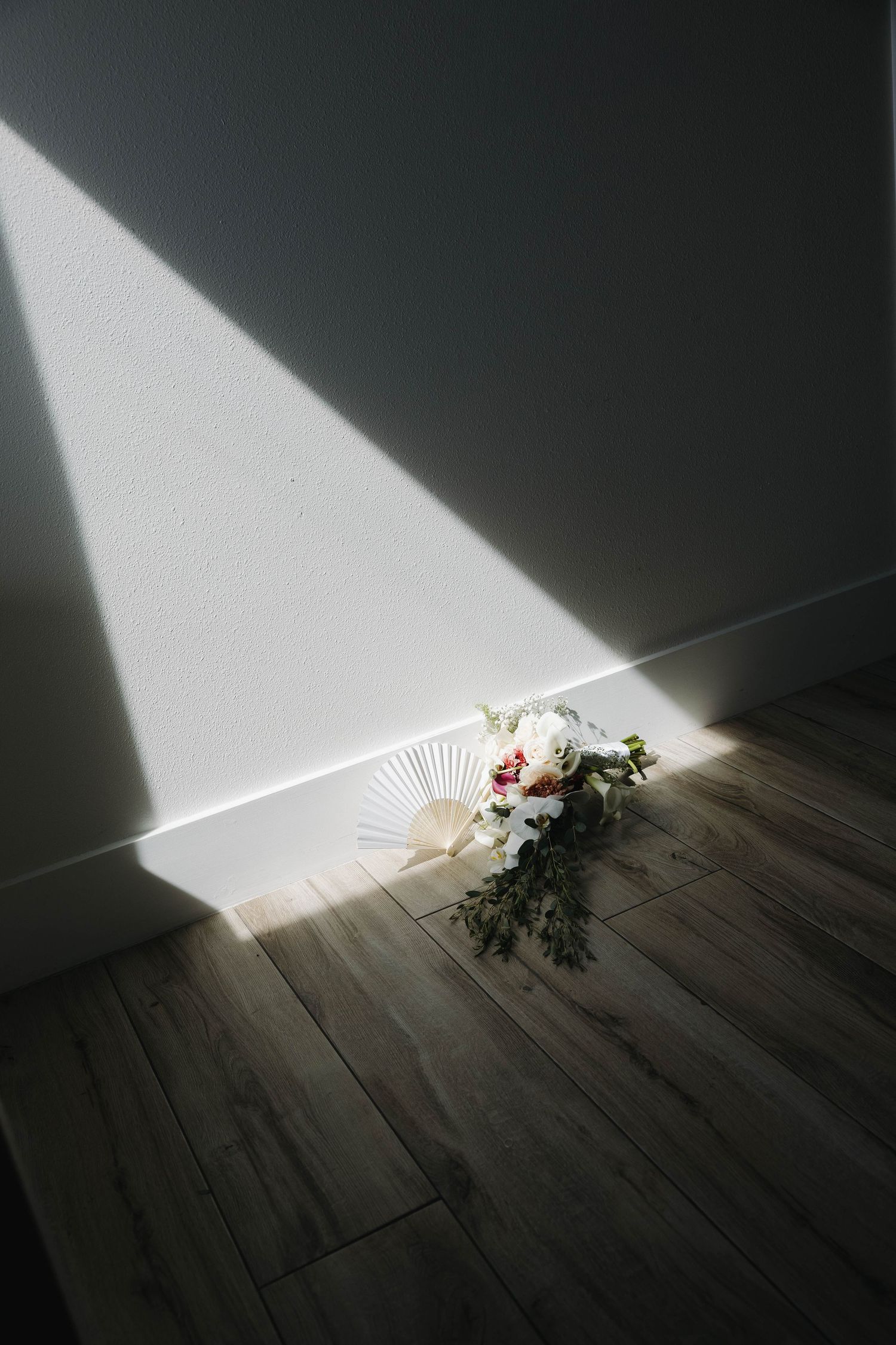 White floral bouquet with paper fan resting on wooden floor in dramatic sunlight beam against white wall.