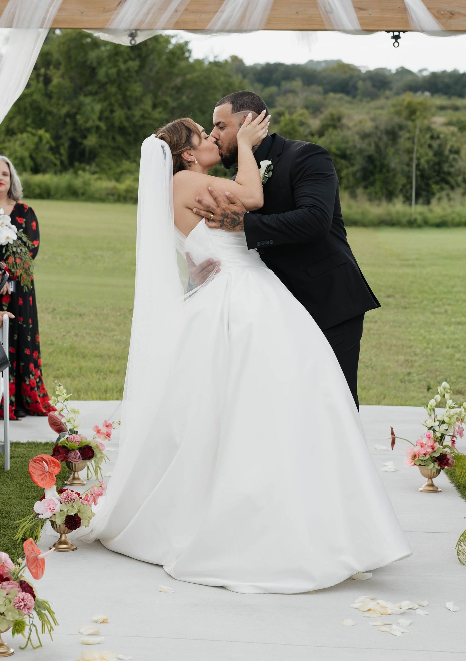 Bride and groom share first kiss at outdoor wedding ceremony with floral arrangements and green field backdrop.