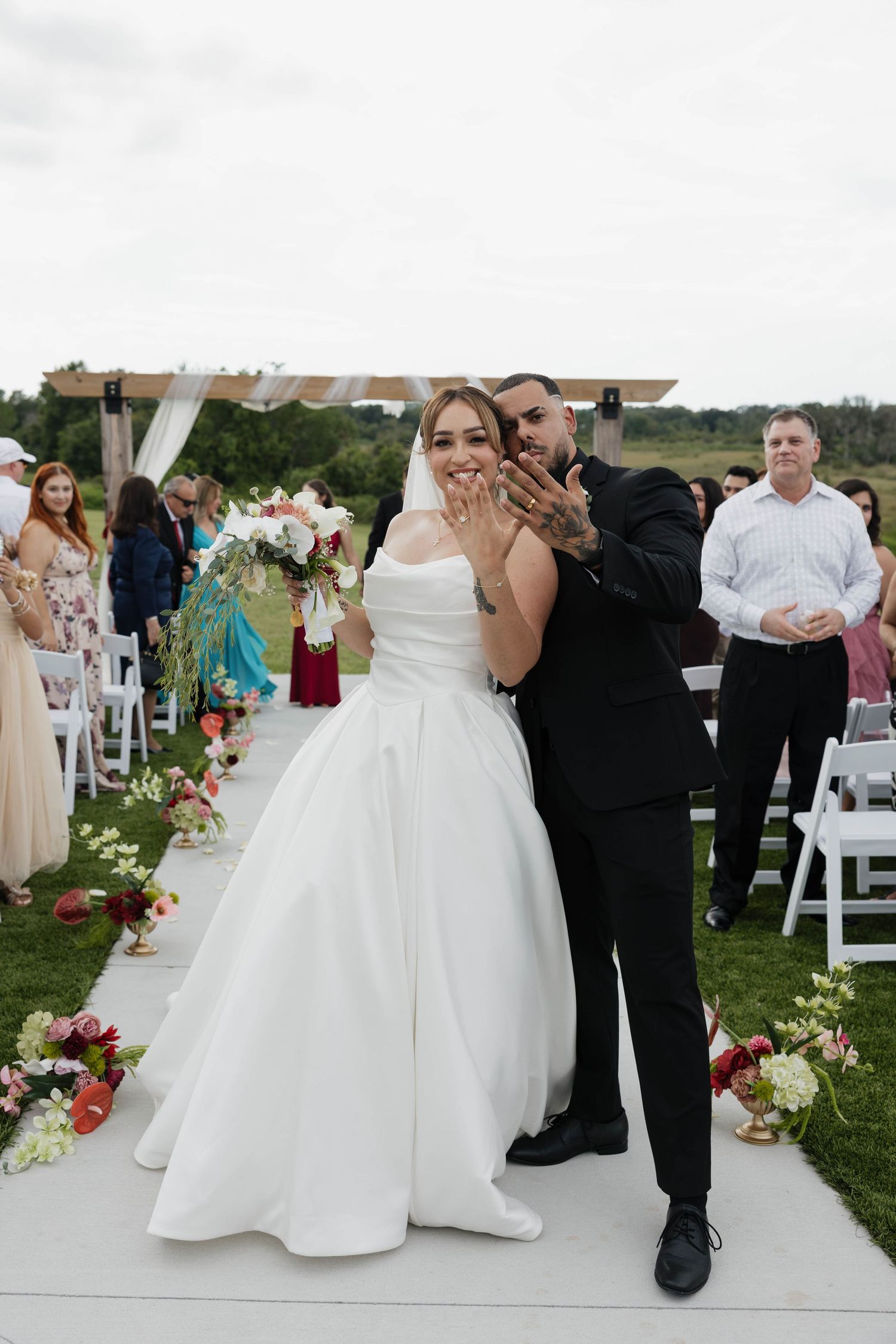 Newlyweds show off wedding rings at outdoor ceremony, bride in white gown holding bouquet, groom in black suit.