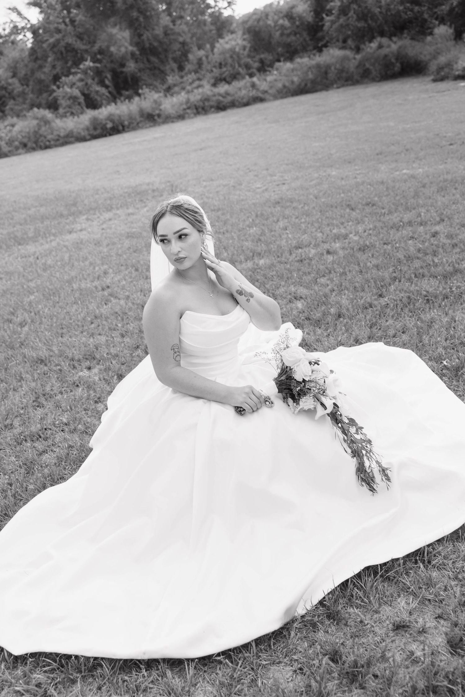 Black and white photo of a bride in a white gown sitting on grass, holding a bouquet, with a veil and serene expression.