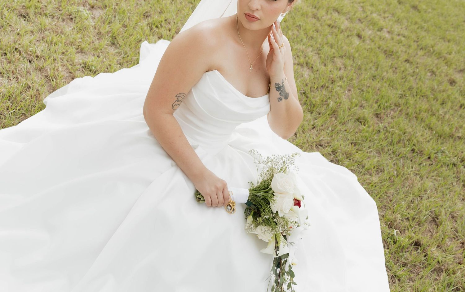 Bride in white strapless ball gown sitting on grass holding a white floral bouquet with red accents.