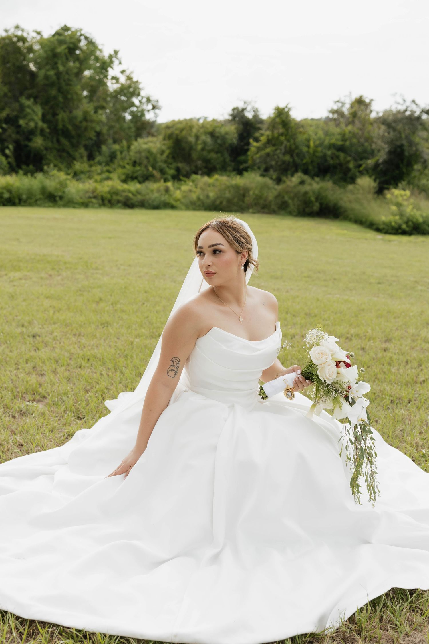 Bride in strapless white ball gown with veil sits on green grass holding white floral bouquet outdoors.