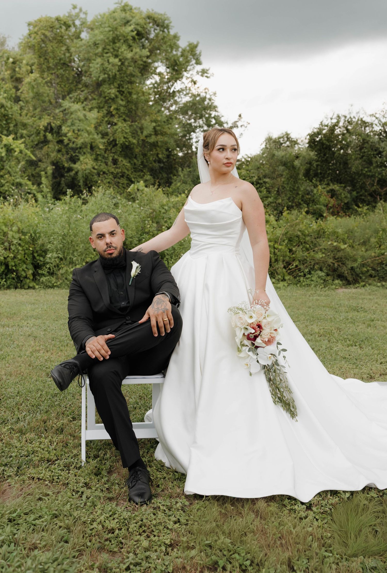 Bride in white strapless gown holding bouquet stands beside groom in black suit seated on chair outdoors.