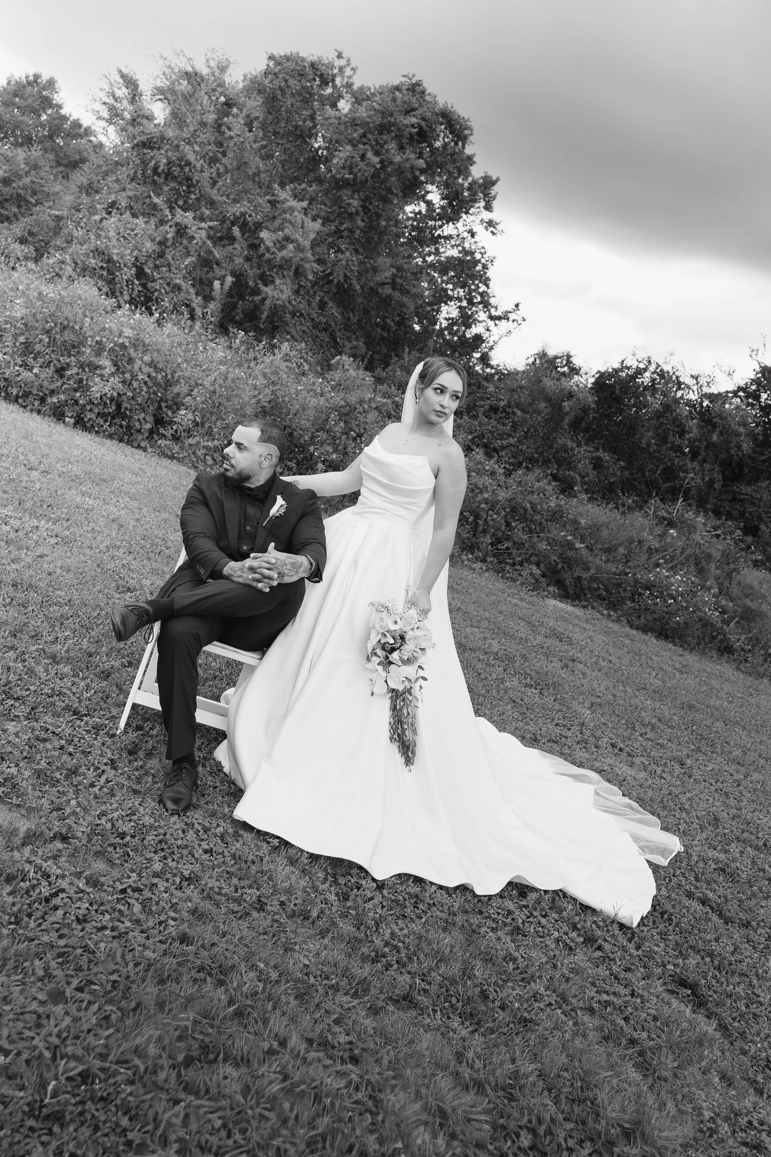 Black and white wedding photo of bride in flowing gown holding bouquet standing beside groom seated on bench outdoors.