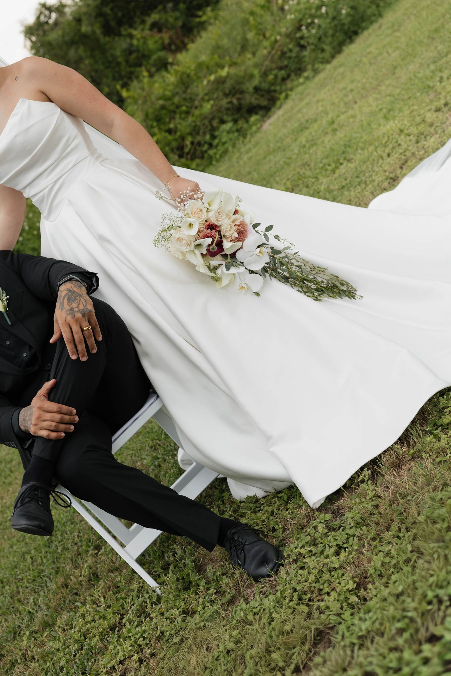 Bride in white gown holds floral bouquet beside groom in black suit seated on chair outdoors on green grass.