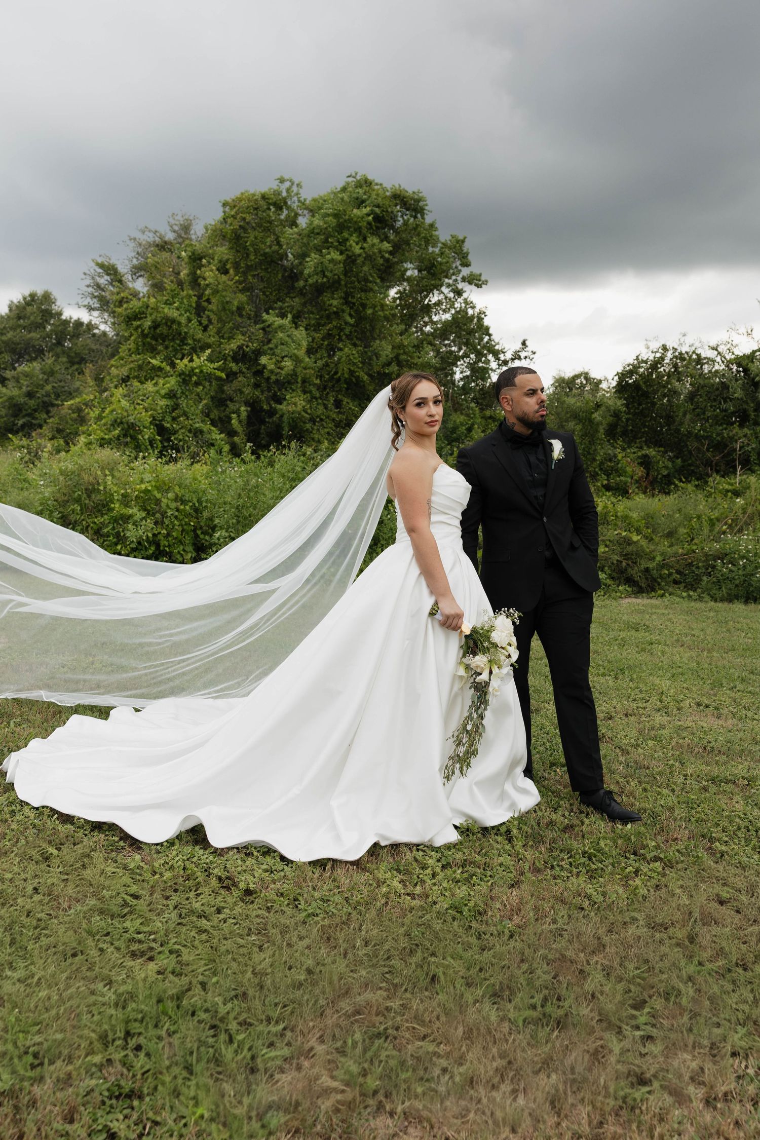 Bride in flowing white gown with veil billowing in wind stands beside groom in black suit on green lawn.