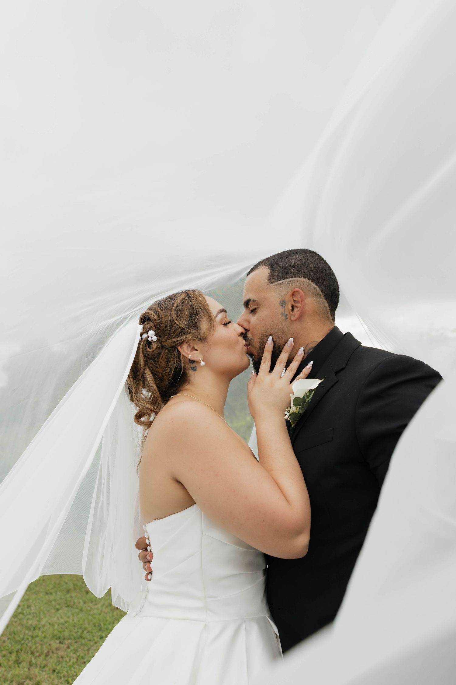 A bride and groom share a romantic kiss under a flowing white veil outdoors on their wedding day.