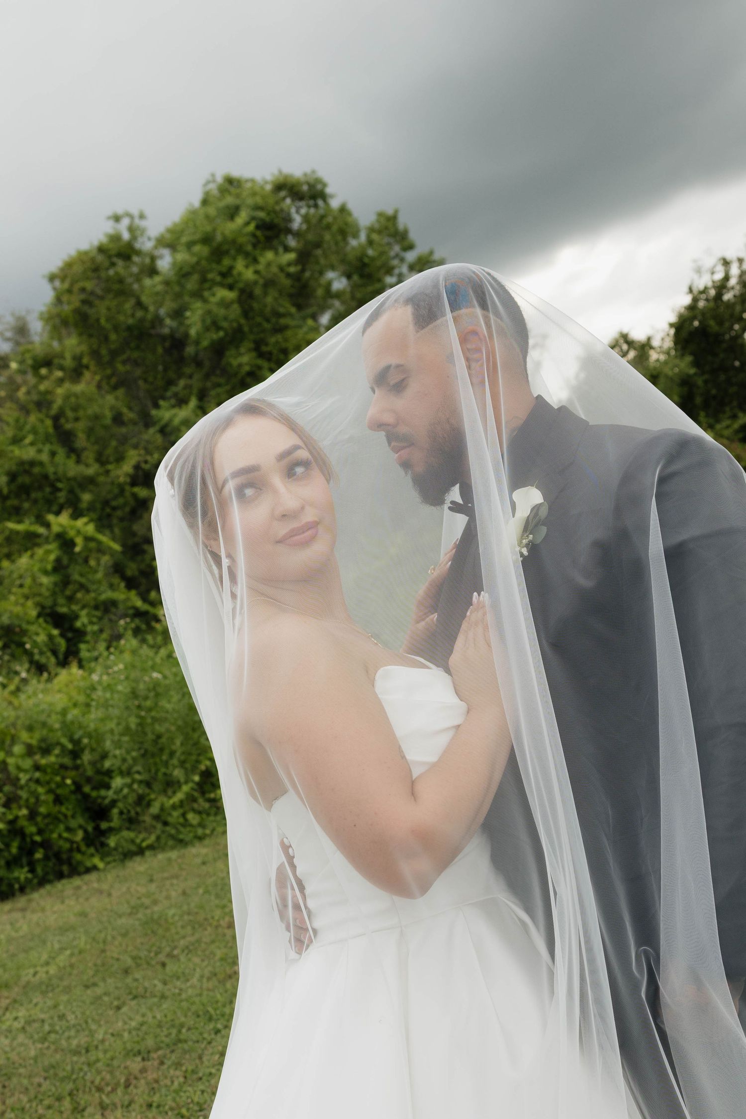 Bride and groom share romantic moment under flowing white veil outdoors with lush green trees and dramatic cloudy sky.