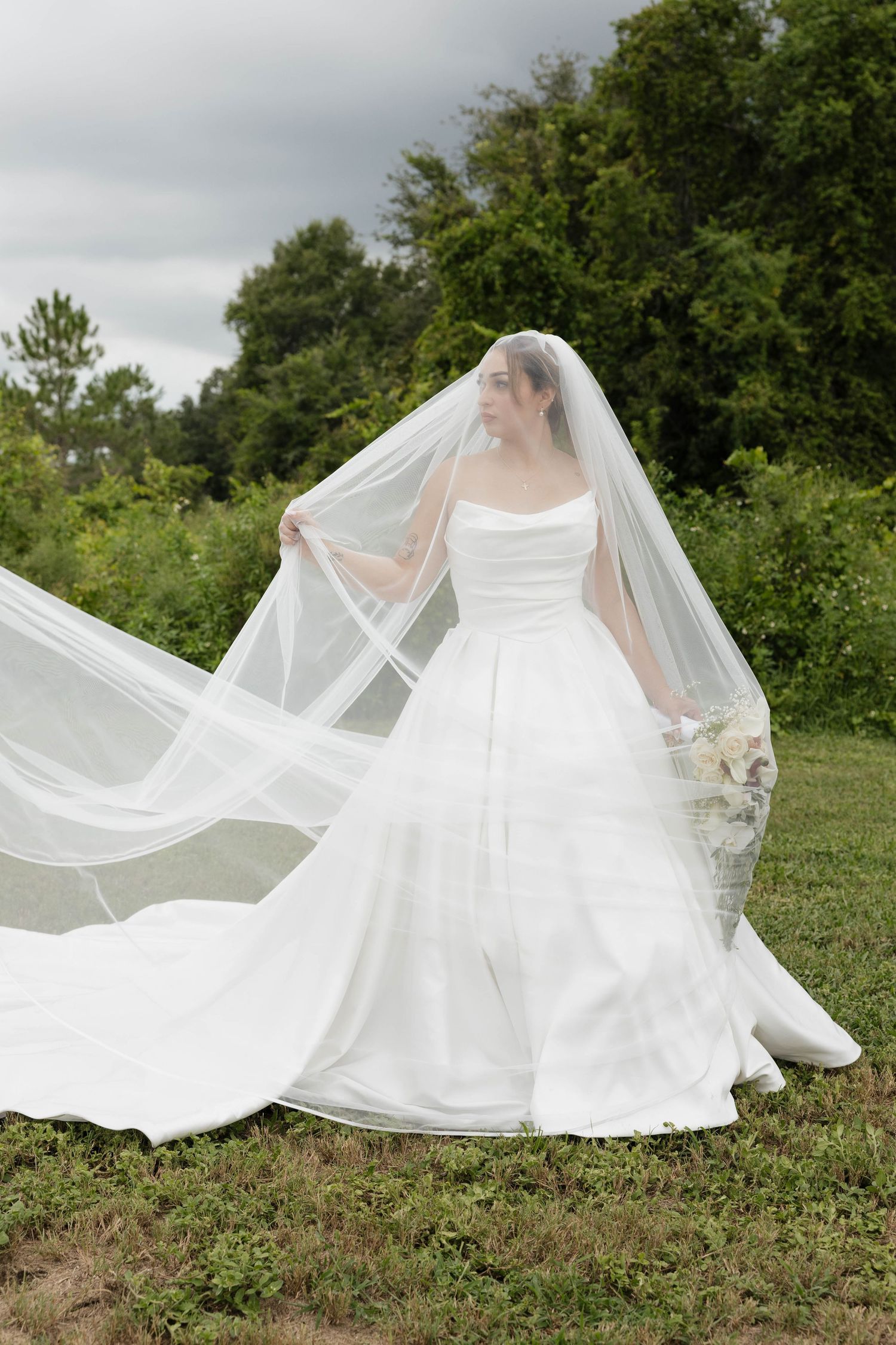Bride in white strapless ball gown with flowing veil outdoors in green field on cloudy day.