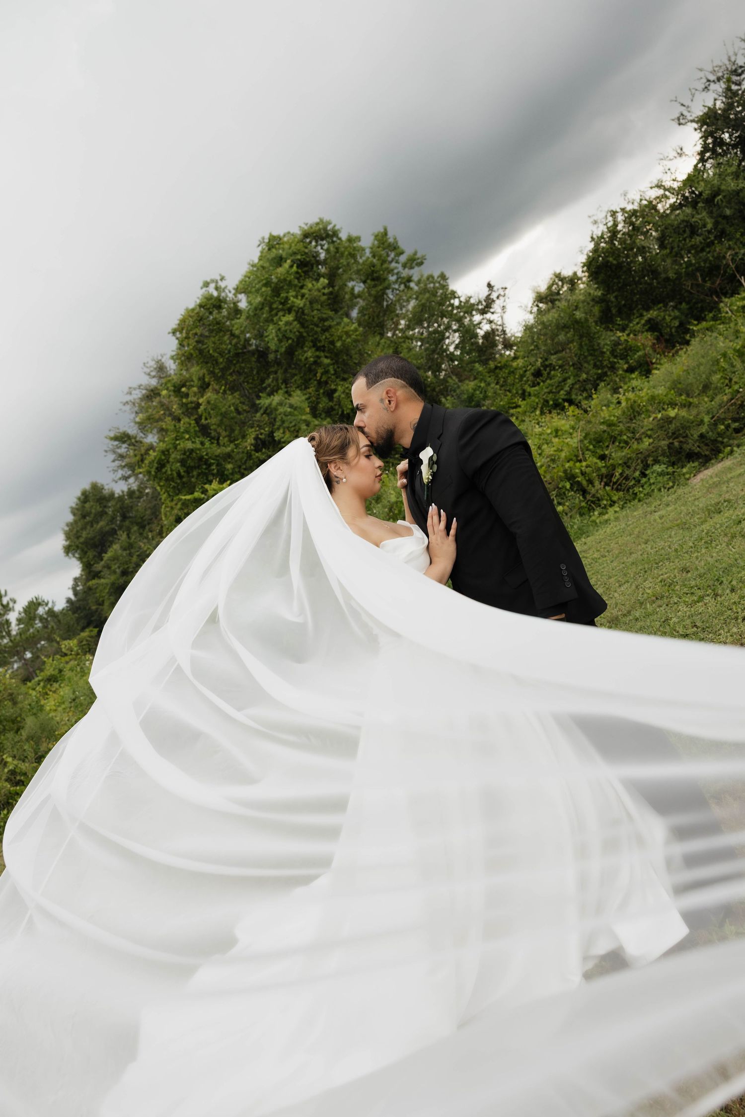 Bride and groom sharing a romantic kiss outdoors, with the bride's flowing white veil billowing in the wind against a dramatic sky.