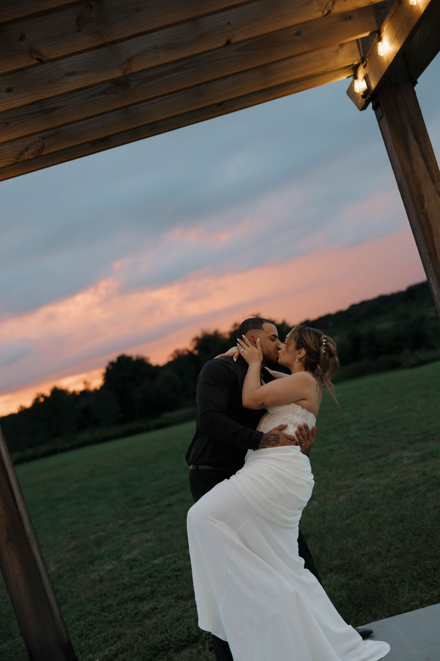 Bride and groom share a romantic kiss under a wooden pergola with string lights at a stunning pink and purple sunset.
