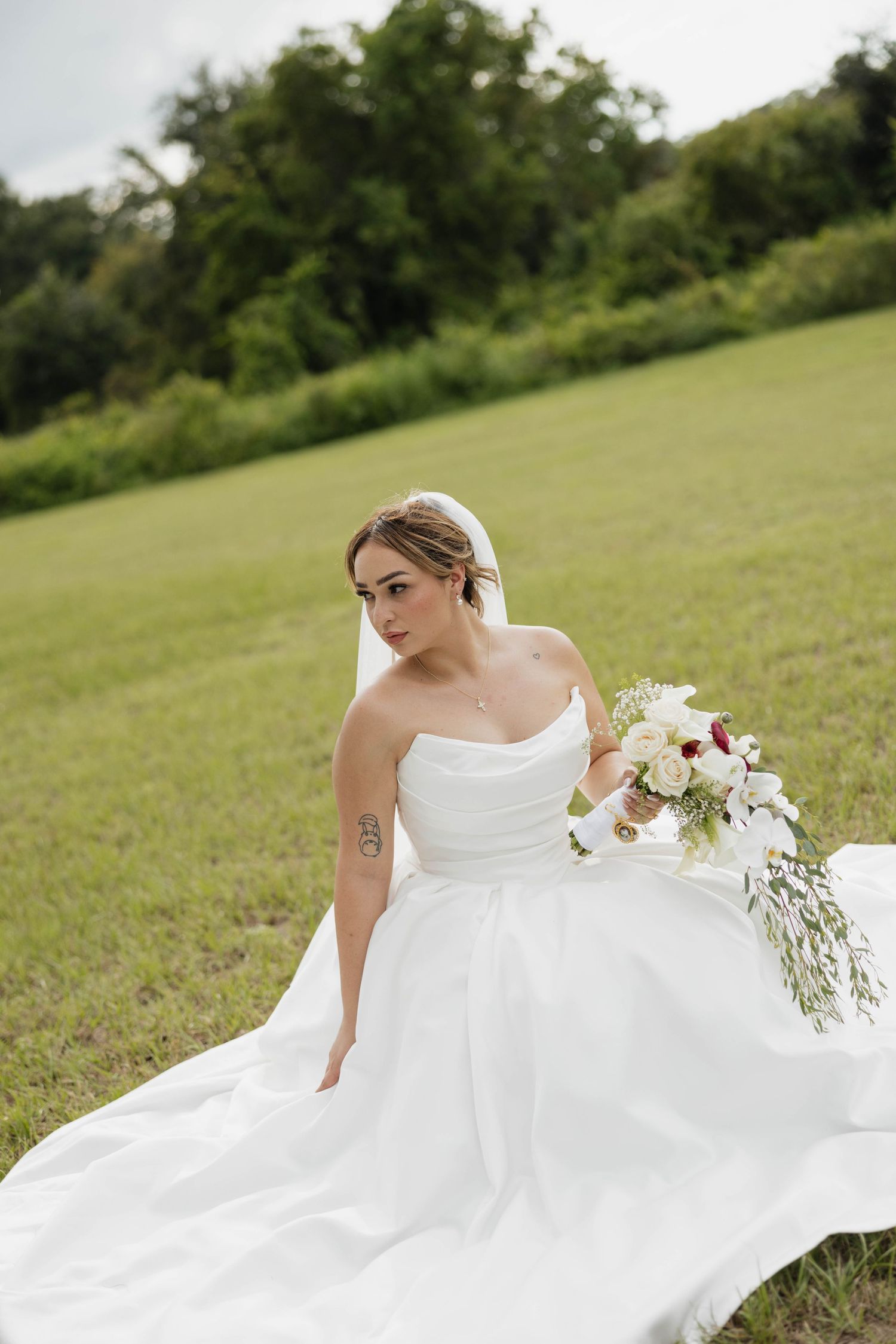Bride in white strapless gown with veil holds floral bouquet while sitting in lush green field on wedding day.