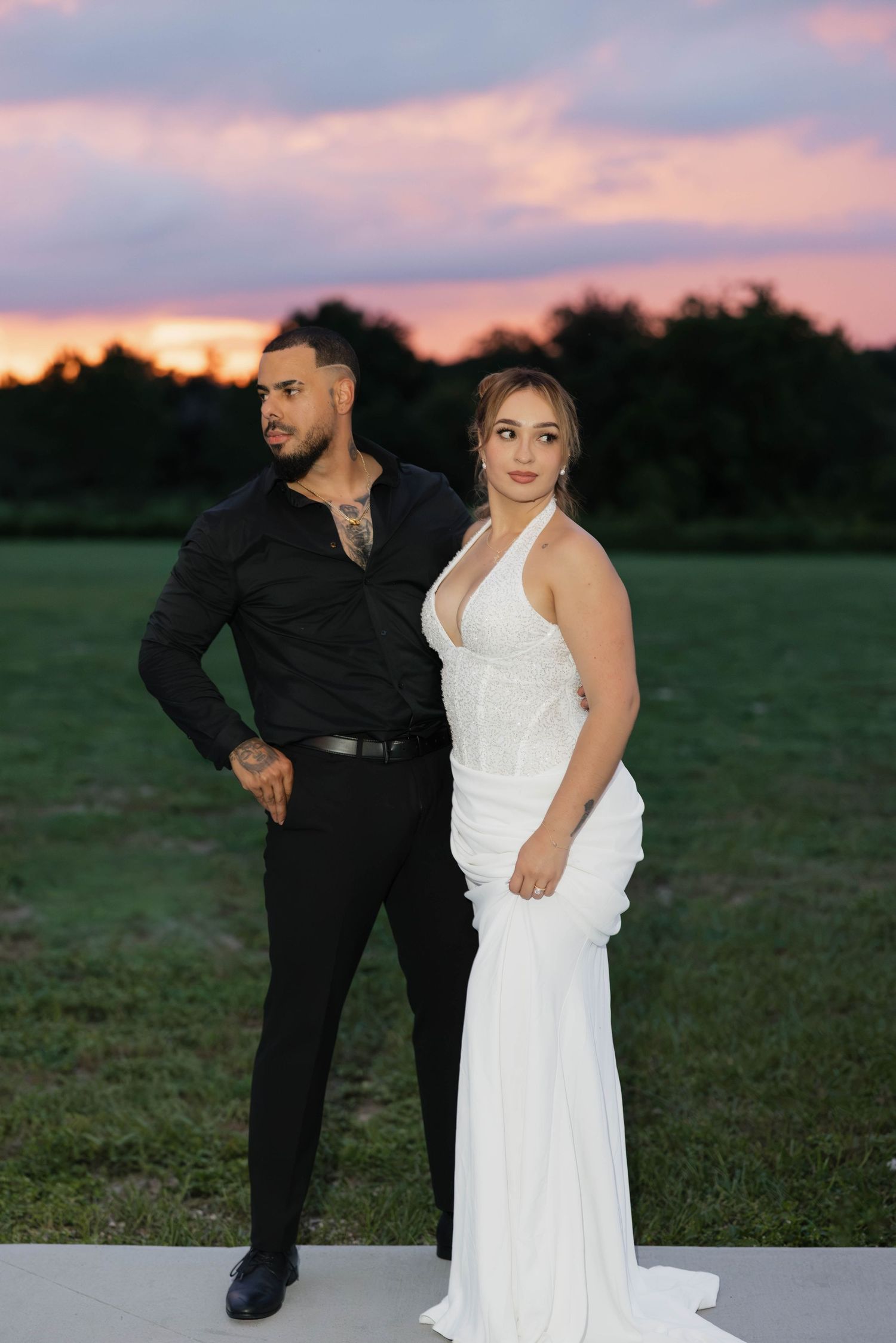 Couple posing at sunset, man in black shirt and pants, woman in white halter wedding gown, colorful sky backdrop.