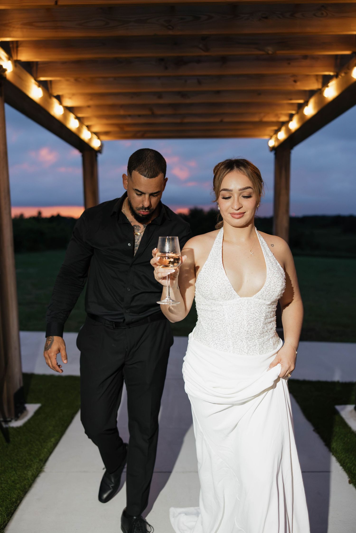 Bride in white halter gown and groom in black suit walk under lit pergola at dusk holding champagne glass.