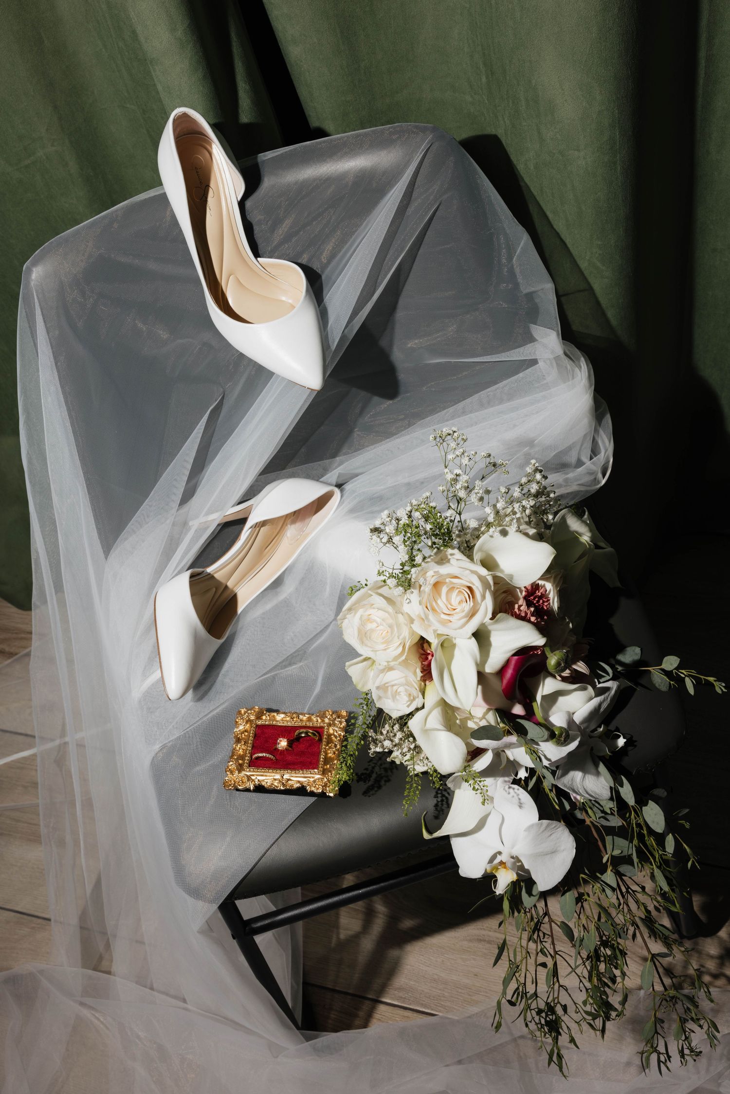 Elegant bridal flat lay with white heels, sheer veil, white rose bouquet, and red velvet ring box on dark stand.