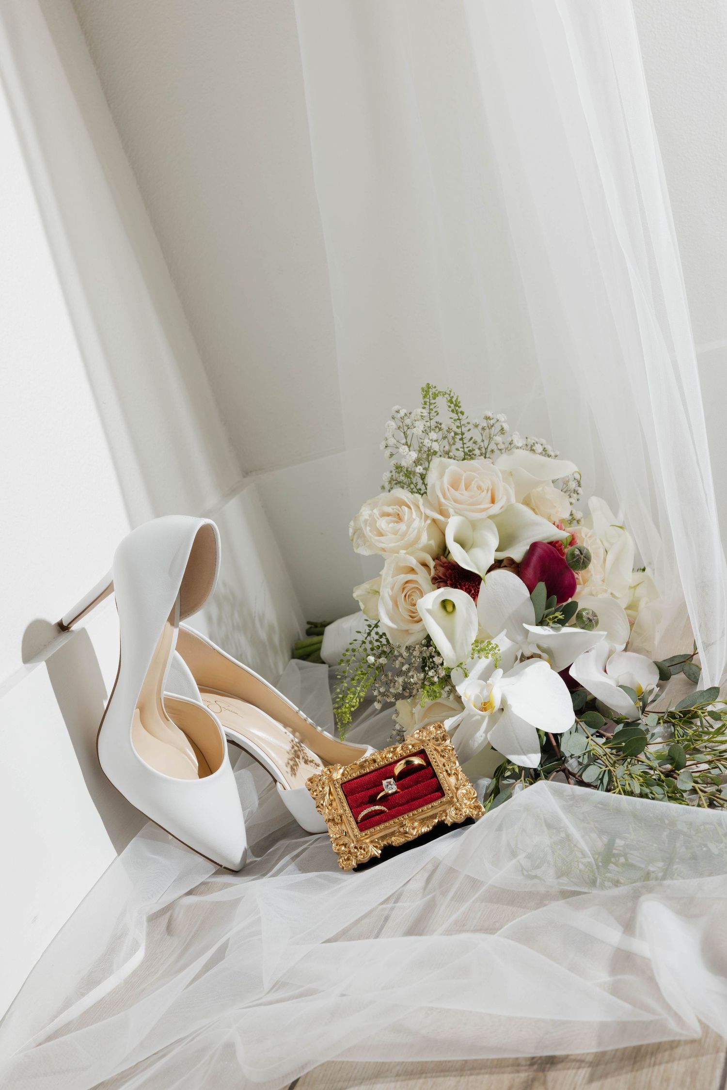 White bridal heels, gold ring box with diamond ring, and elegant bouquet of roses and orchids on soft white fabric.