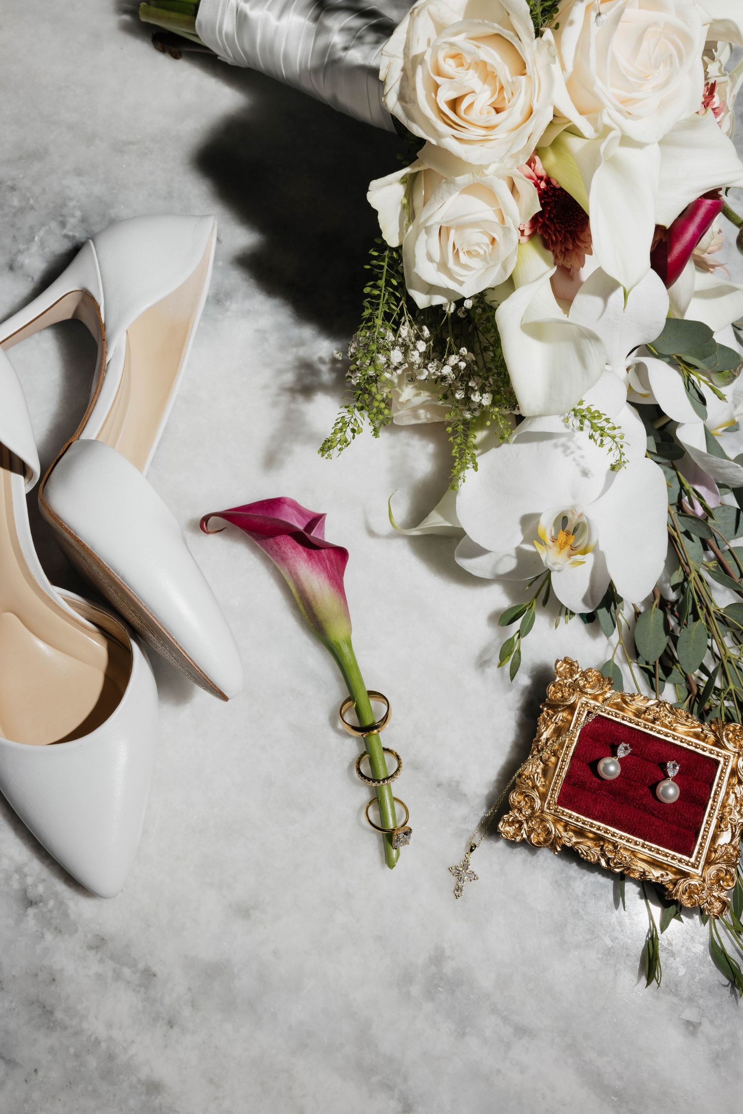 Elegant bridal flat lay with white heels, wedding rings on calla lily, pearl earrings in gold frame, and white rose bouquet.