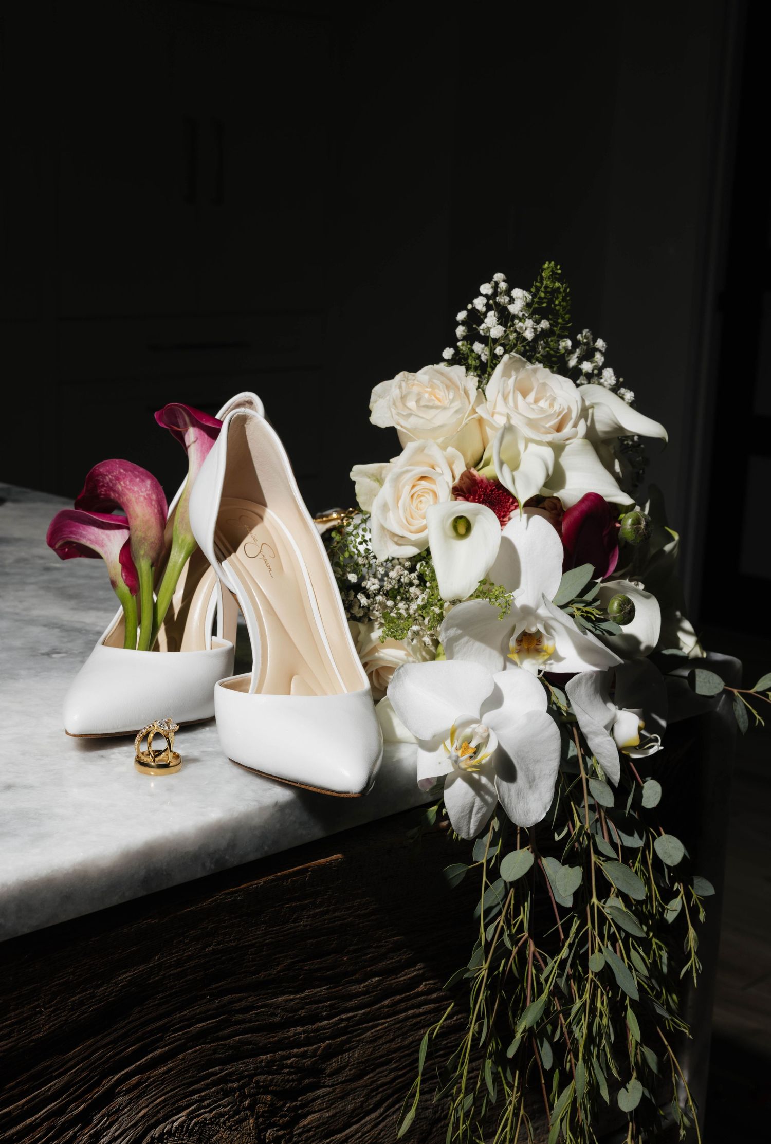 White bridal heels and wedding rings beside an elegant bouquet of white orchids, cream roses, and deep purple calla lilies.