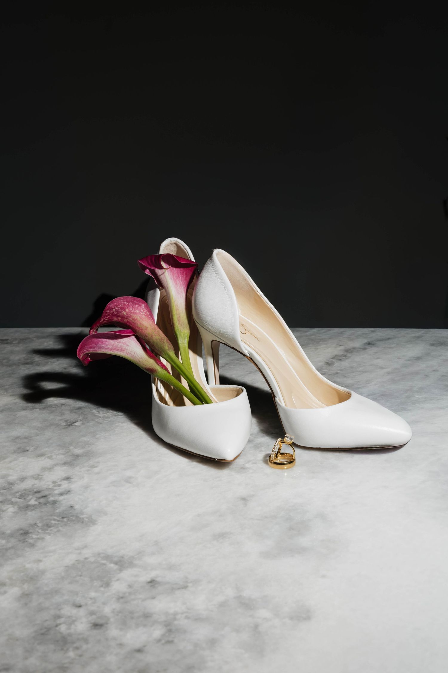 White bridal heels with pink calla lilies on marble surface against dark background.