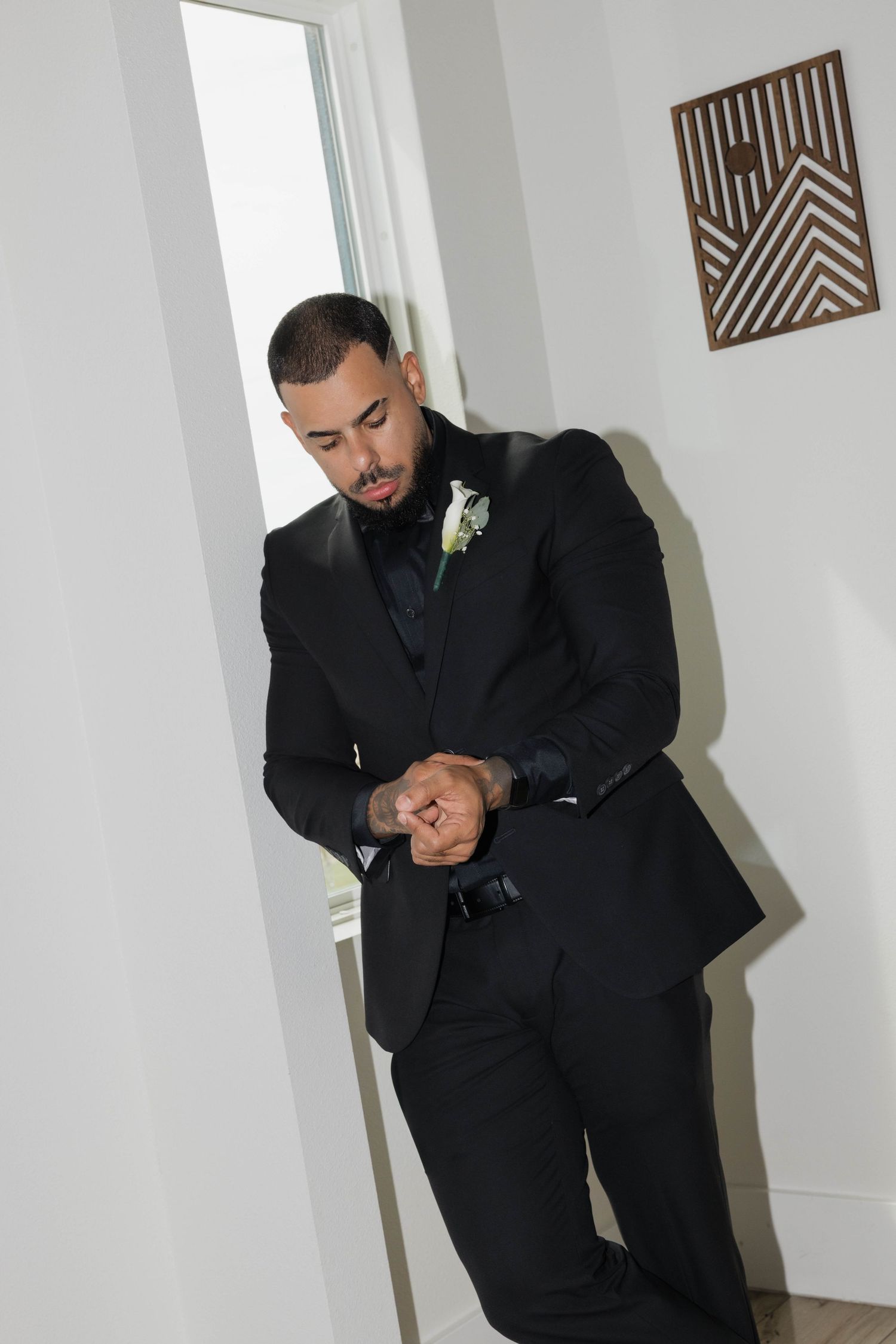 Groom in black tuxedo with boutonniere adjusting cufflinks while leaning against a wall indoors.
