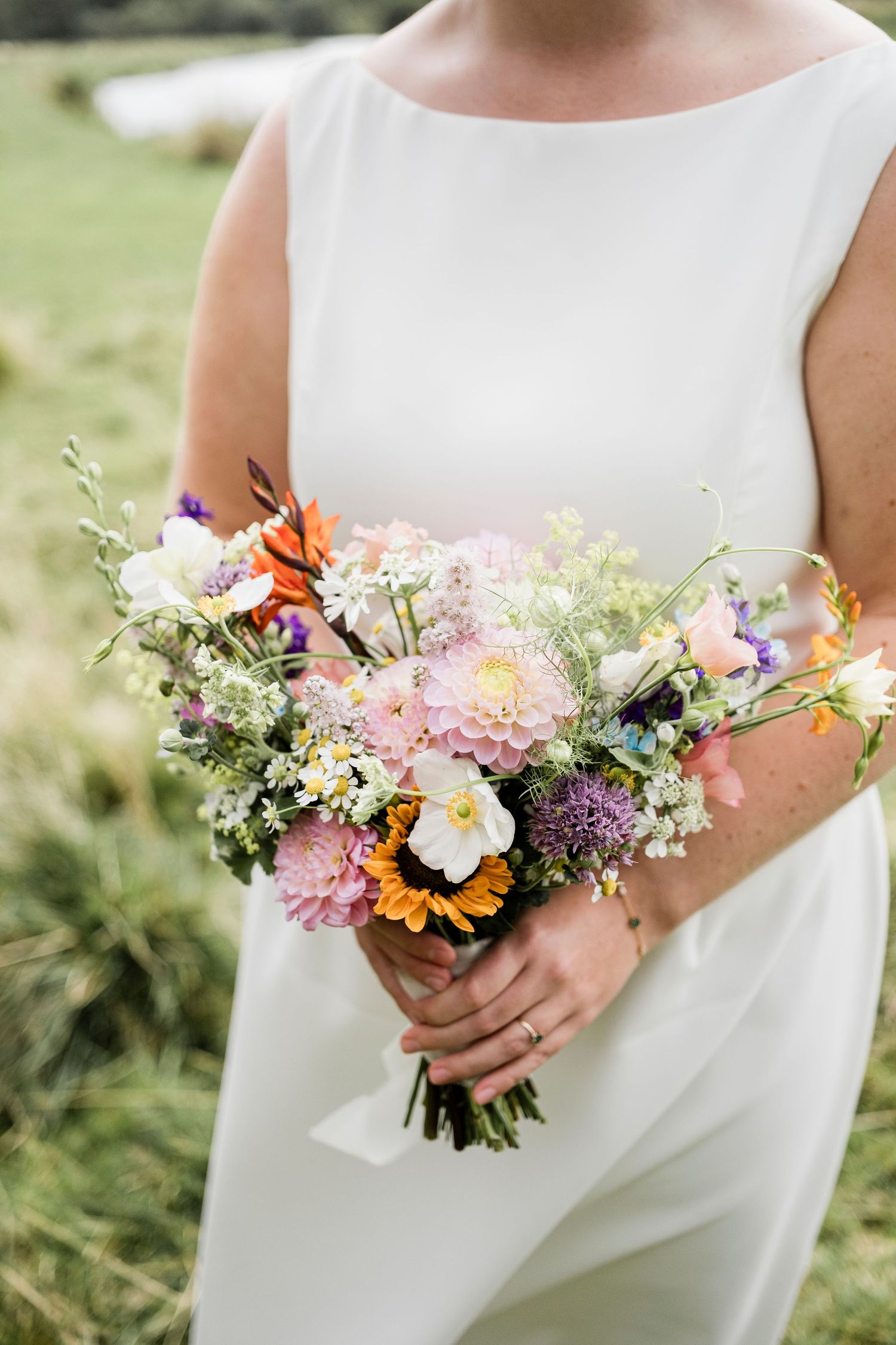 Bride in white dress holding a colorful wildflower wedding bouquet with pink, orange, and purple blooms outdoors.