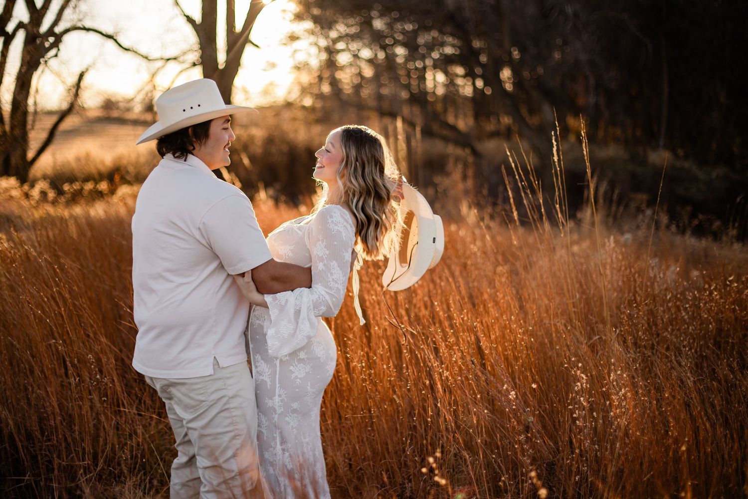 Couple in white outfits embracing in golden tall grass field at sunset, man wearing cowboy hat.