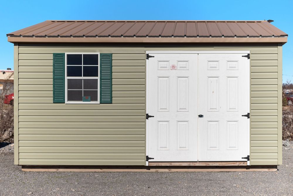 Tan vinyl storage shed with brown metal roof, green shutters, white double doors, and a single window.