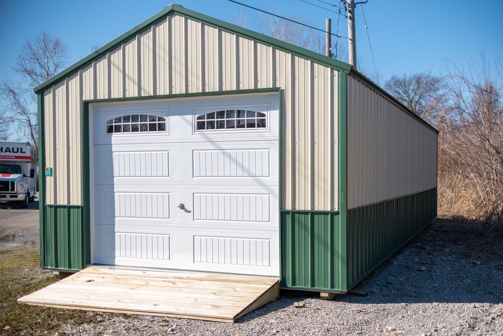 Green and beige metal garage building with white overhead door and wooden ramp on gravel lot.