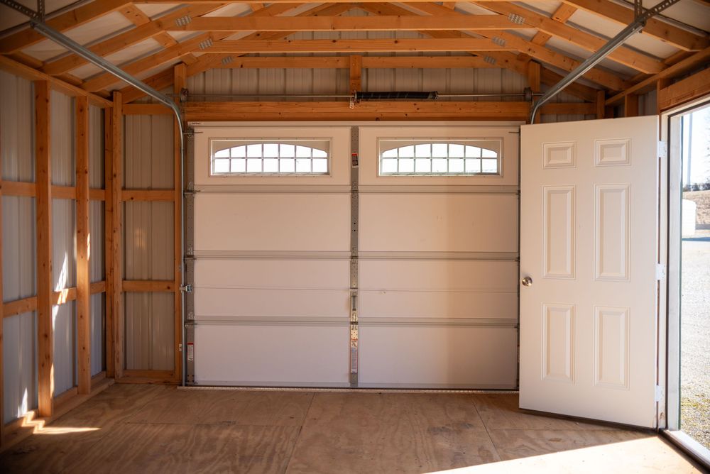Interior view of a wooden garage with white double garage door and side entry door on a dirt floor.