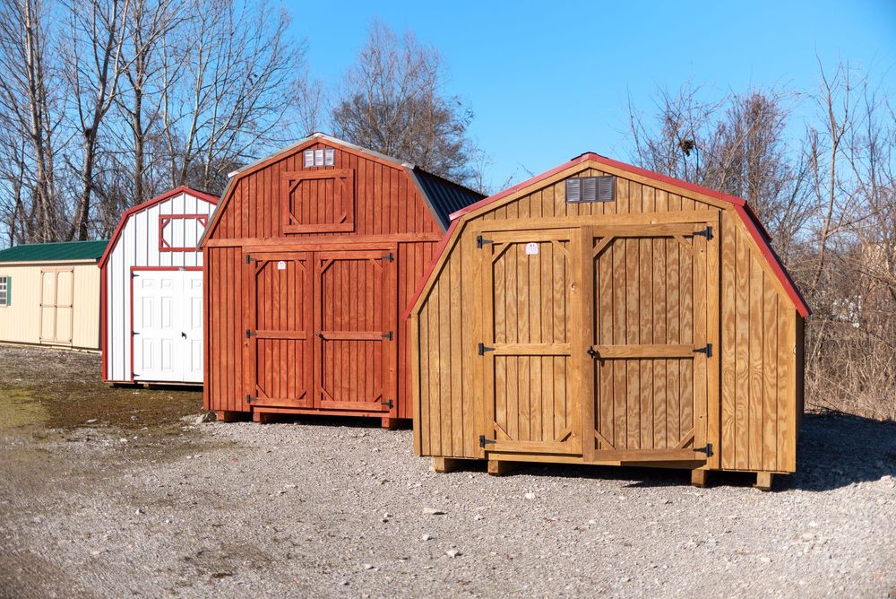 Three wooden storage sheds in red and natural wood tones displayed outdoors on gravel with trees in the background.