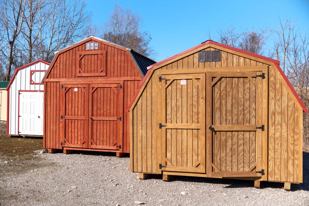 Two wooden storage sheds with barn-style roofs displayed outdoors, one stained red and one natural wood tone.