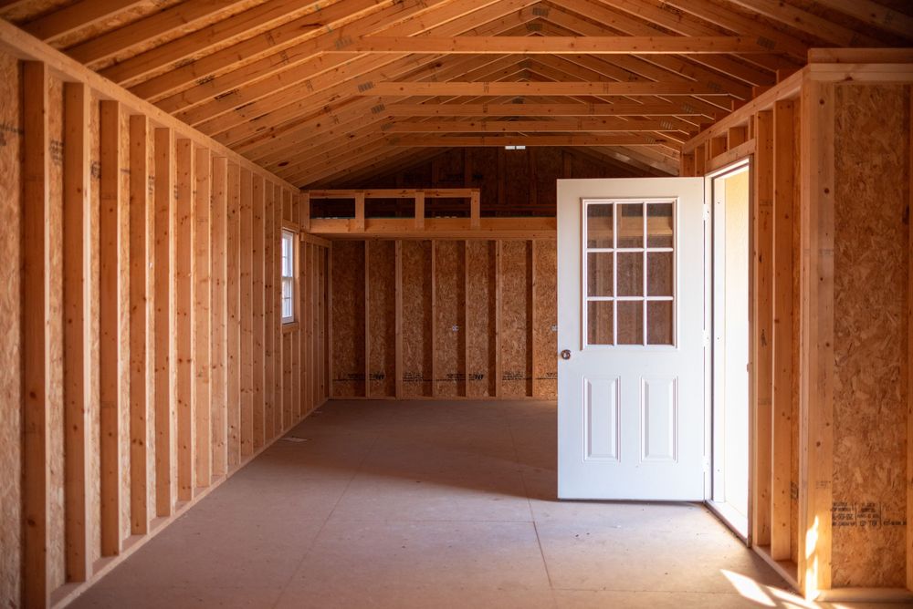 Interior of a wooden storage shed with vaulted ceiling, white door, and unfinished pine walls on a concrete floor.