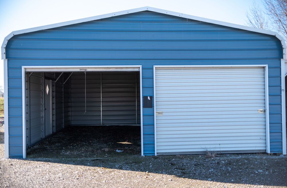 Blue metal garage building with two bays, one open and one with a white roll-up door, on gravel.