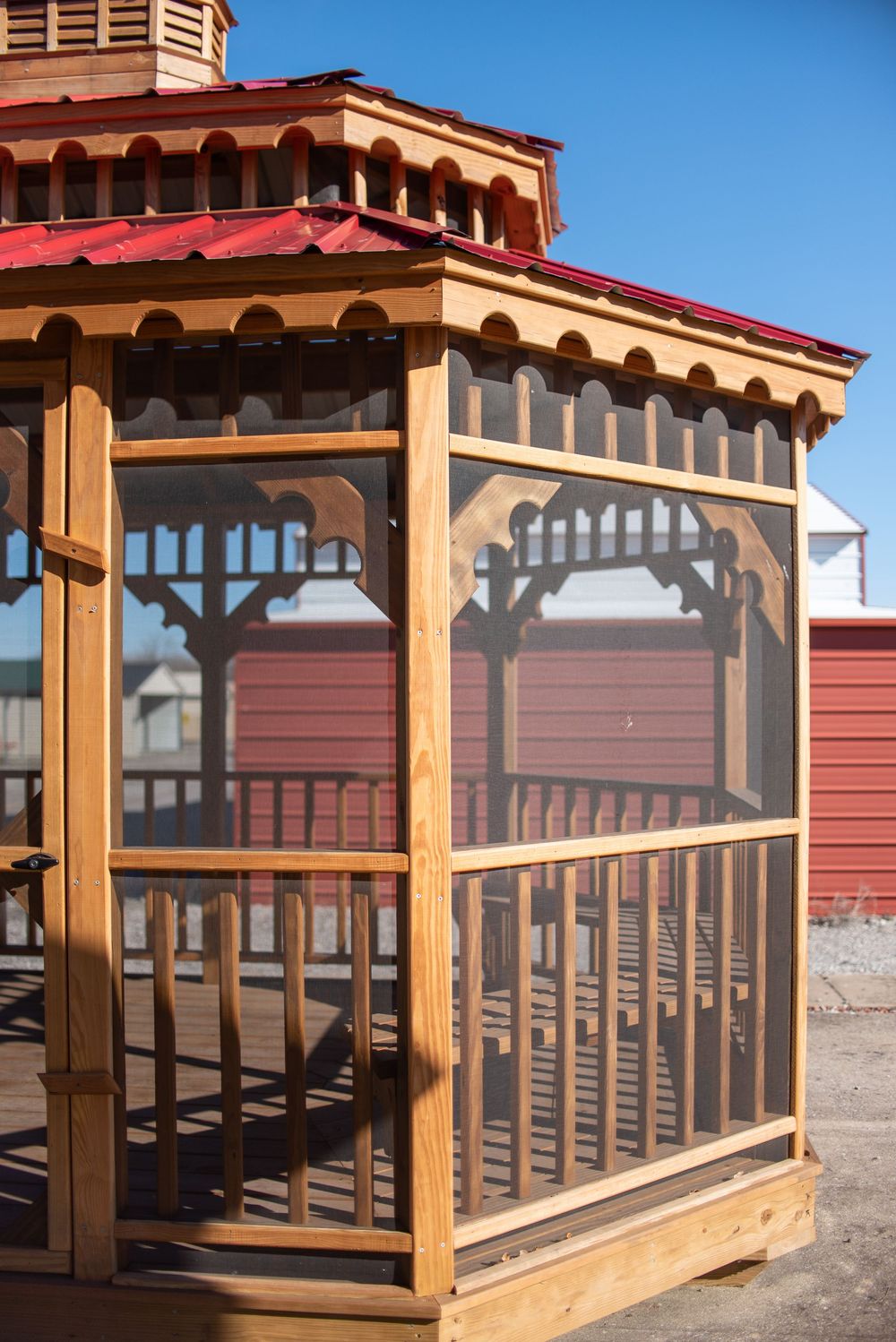 Wooden gazebo with red roof and screened walls displayed outdoors on a sunny day.