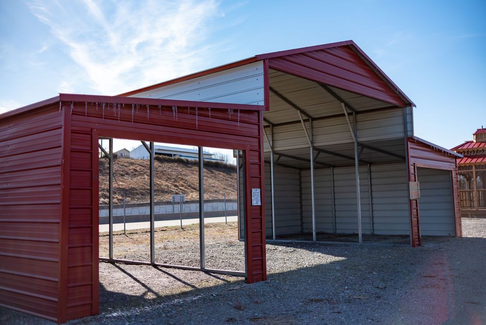 Red metal carport with open sides and attached storage unit on gravel, set against a clear blue sky.